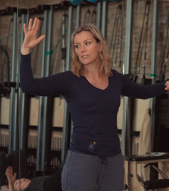 Helen, founder and teacher at The Movement Retreat, leading a class in a rustic brick-walled studio. She is wearing a dark long-sleeve top and grey joggers, with her arms outstretched as she guides her students. Pilates equipment and class participants can be seen in the background.